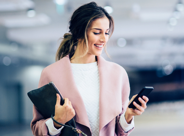 Geschäftsfrau mit dunklen Haaren und rosa Mantel, hält Handy in der Hand und Laptop unterm Arm