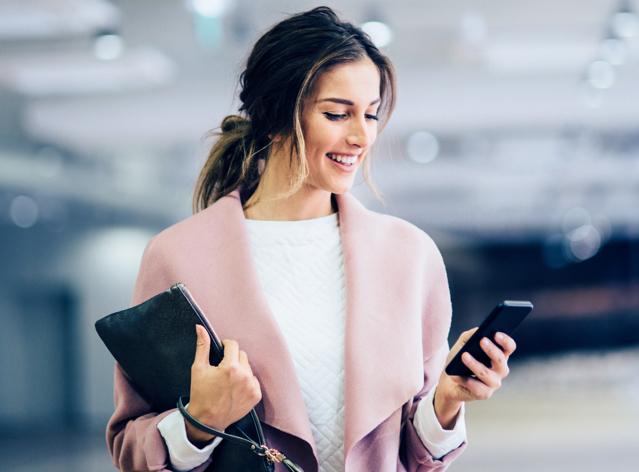 Frau mit Mobiltelefon Geschäftsfrau mit dunklen Haaren und rosa Mantel, hält Handy in der Hand und Laptop unterm Arm