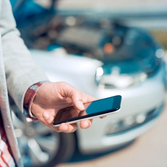 Legerer Geschäftsmann steht beim parkenden Auto und hält ein Handy in der Hand