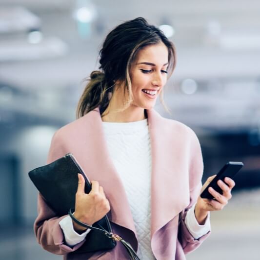 Geschäftsfrau mit dunklen Haaren und rosa Mantel, hält Handy in der Hand und Laptop unterm Arm