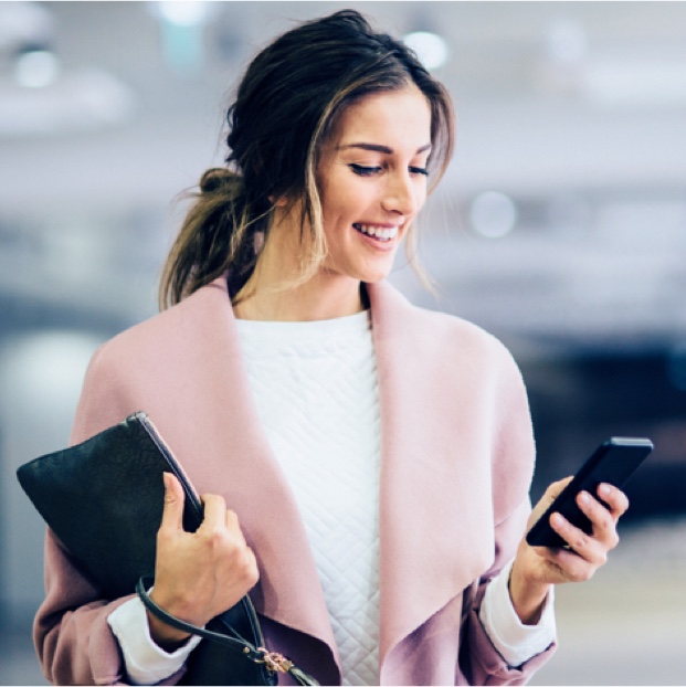 Frau mit Mobiltelefon in der Hand Es ist eine Geschäftsfrau mit dunklen Haaren und rosa Mantel zu sehen. Sie hält ein Handy in der linken Hand und einen Laptop unter dem rechten Arm.