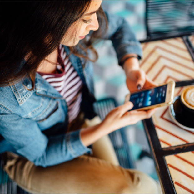 Es ist eine Frau in Freizeitkleidung zu sehen. Sie sitzt in einem Café und blickt auf ihr Mobiltelefon.