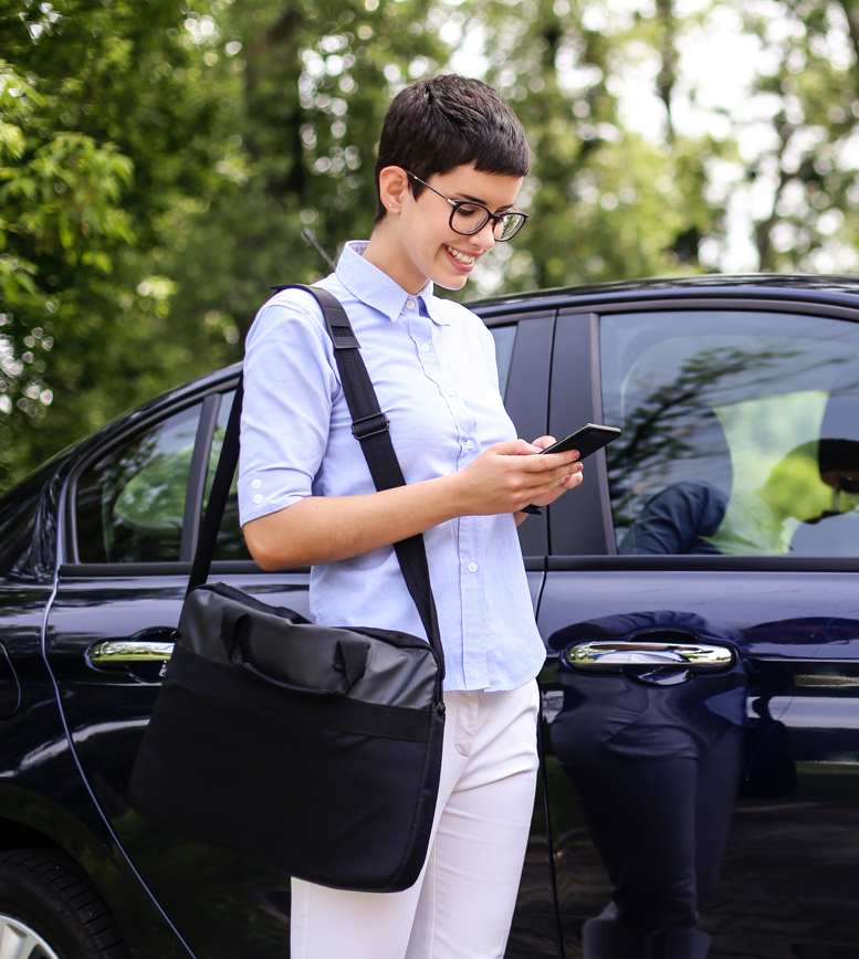 Frau mit Smartphone vor Auto im Park Es ist eine Frau mit kurzen braunen Haaren und einer Brille zu sehen. Sie hält ein Handy in der Hand und trägt eine Laptop-Umhängetasche. Sie steht vor einem schwarzen PKW.